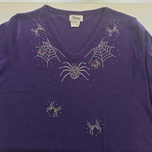 Peut inclure: Un t-shirt violet &agrave; manches longues et col en V orn&eacute; d'araign&eacute;es et de toiles d'araign&eacute;es en strass. Les araign&eacute;es ont des accents rouges, blancs et verts. Le t-shirt porte une &eacute;tiquette "Distinct".