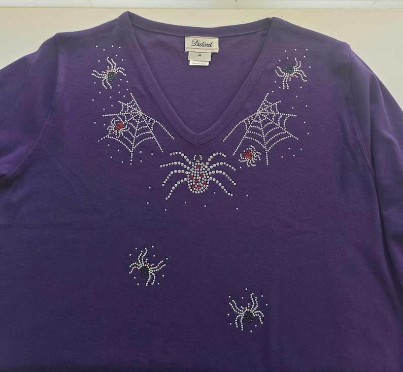 Peut inclure: Un t-shirt violet &agrave; manches longues et col en V orn&eacute; d'araign&eacute;es et de toiles d'araign&eacute;es en strass. Les araign&eacute;es ont des accents rouges, blancs et verts. Le t-shirt porte une &eacute;tiquette "Distinct".