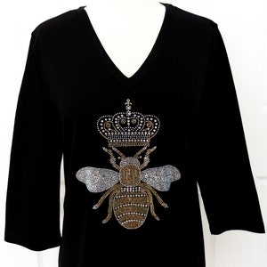 May include: Black t-shirt featuring a bee design adorned with rhinestones, wearing a crown. The bee and crown are made of gold and silver rhinestones.