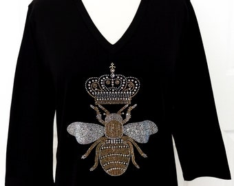 Puede incluir: Camiseta negra con un diseño de abeja de pedrería que lleva una corona. La abeja y la corona están hechas de pedrería dorada y plateada.