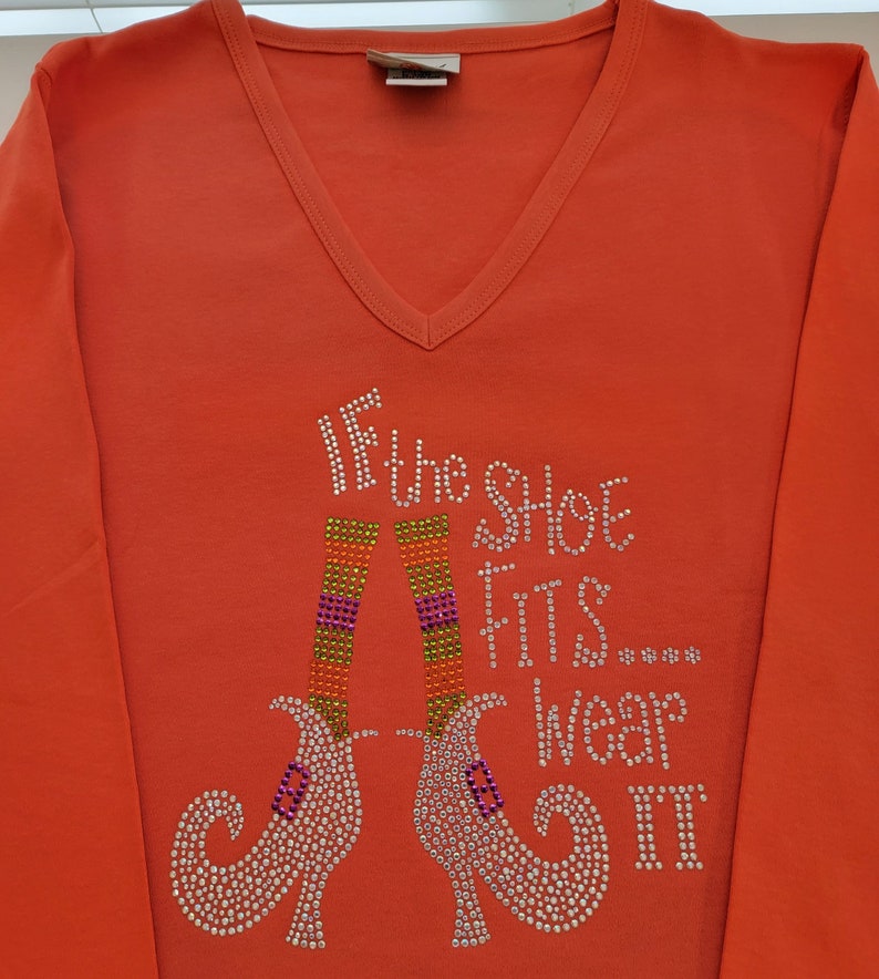 May include: Orange long-sleeved shirt with a sparkly design of two witch's shoes and the text "If the shoe fits... wear it".