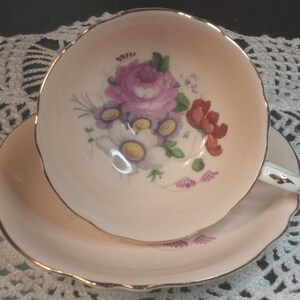 Può includere: Un set vintage di tazza e piattino da tè. La tazza e il piattino rosa pallido hanno bordi smerlati e finiture dorate. La tazza presenta un motivo floreale con fiori viola, rossi e bianchi. Il manico è bianco con dettagli dorati.