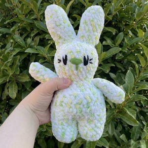 May include: A handmade, plush bunny rabbit in white with green and yellow accents. The bunny has long ears, black eyes, and a green nose. The soft toy is held in front of a green leafy background.