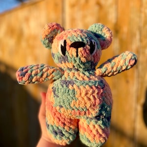 May include: A handmade crochet teddy bear in pastel colors of orange, green, blue, and pink. The bear has a black stitched face and is held in a person's hand. The bear's arms and legs are extended.