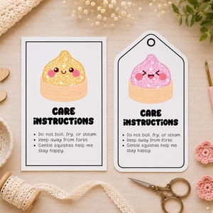 Op de afbeelding: Twee witte kaarten, een rechthoekig en een in labelvorm, met onderhoudsinstructies en illustraties van gouden en roze glitter-dumplings. De tekst luidt: CARE INSTRUCTIONS, Do not boil, fry, or steam, Keep away from forks, Gentle squishes help me stay happy.