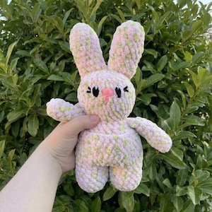 May include: A handmade crochet bunny in soft pink with green and yellow accents. The plush toy has long ears, a pink nose, and black embroidered eyes. The bunny is a cuddly, whimsical creation.