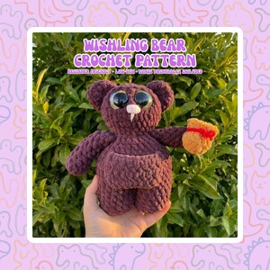May include: A crocheted bear in shades of purple, holding a small, tan pouch with a red ribbon. The bear has large, expressive eyes and a white snout. Text on the image reads "Wishing Bear Crochet Pattern".