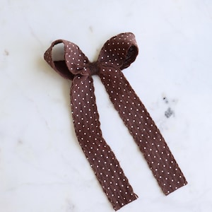 May include: A brown fabric hair bow featuring a repeating pattern of small white polka dots. The bow has two long, dangling ribbon tails with frayed, raw edges and a central knot detail that secures the loops and tails together in a classic shape.