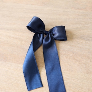 May include: A navy blue satin bow with long, flowing tails. The bow is tied in the center and sits on a light wooden surface. The ribbon has a slight sheen.