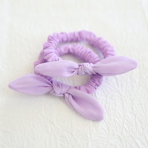 May include: Two lavender fabric hair scrunchies with bow details.