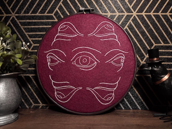 A finished embroidery hoop crawling with surreal, hyper-detailed eyes — hand-stitched fiber art that's genuinely unsettling and beautiful at once.