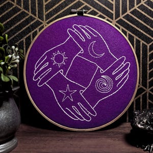 May include: A purple embroidery hoop features four hands in a circle, each with a different symbol: sun, moon, star, and spiral. The design is stitched in white thread on a purple background, set in a wooden hoop. The background is a dark geometric pattern.