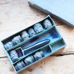 May include: A vintage stamp set in a light blue box. The set includes several small, cylindrical stamps with various embossed designs, a dark handle, and a small spool of purple thread. The box is open, revealing the stamps and handle.