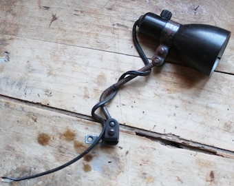 May include: A vintage black spotlight with a metal bracket and a twisted wire. The spotlight has a small adjustment knob and a simple design. The electrical wire has exposed ends.