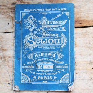 May include: A vintage blue album cover with white ornate text and illustrations. The cover features the text "OUVRAGES DAMES MAISON SAJOU" and "ALBUMS". The cover also includes the text "74-Boulevard Sebastopol-74 PARIS".