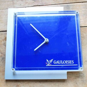 May include: A blue clock with a silver frame and hands. The clock face has the word "GAULOISES" printed on it.