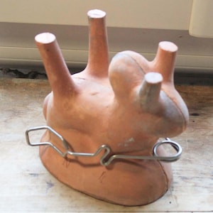 Könnte beinhalten: Ein terrakottafarbenes Dentalabdruckmodell mit vier zylindrischen Vorsprüngen und einem Metalldrahtbauteil. Das Modell hat eine abgerundete Basis und dient zur Aufnahme von Dentalabdrücken.