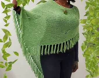 Peut inclure: Un poncho tricoté vert clair avec des franges. Le poncho est porté sur un haut noir à manches longues et un pantalon noir. Le modèle sourit et porte un collier. Des feuilles vertes sont en arrière-plan.
