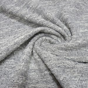 Può includere: Primo piano di un tessuto strutturato con un motivo ondulato. Il tessuto è un mix di grigio e bianco, creando un aspetto screziato. Il materiale è piegato, mostrando la trama e la profondità della tessitura. Ideale per abbigliamento o arredamento.