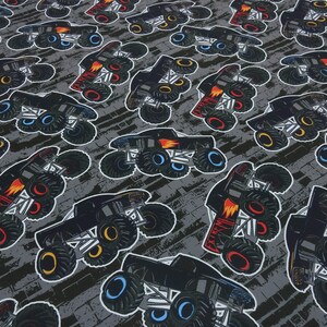 Könnte beinhalten: Ein grauer und schwarzer Stoff mit einem sich wiederholenden Muster von Monstertrucks. Die Trucks sind schwarz mit roten Flammen und haben große, bunte Räder.