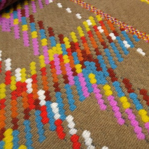 Puede incluir: Primer plano de una alfombra tejida marr&oacute;n con rayas de colores rosa, azul, amarillo, naranja y rojo. Las rayas est&aacute;n hechas de peque&ntilde;as bolas redondas y peludas.