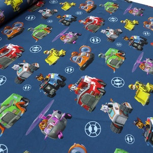Könnte beinhalten: Ein blauer Stoff mit einem sich wiederholenden Muster aus Cartoon-Hunden, die verschiedene Fahrzeuge fahren. Die Hunde tragen verschiedene farbige Uniformen und fahren ein Feuerwehrauto, ein Polizeiauto, einen Hubschrauber, ein Baufahrzeug und einen Rettungswagen. Der Hintergrund ist einfarbig blau.