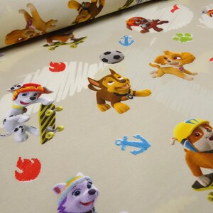 Könnte beinhalten: Ein beigefarbener Stoff mit einem sich wiederholenden Muster aus Cartoon-Hunden aus der Zeichentrickserie Paw Patrol. Die Hunde sind in verschiedenen Posen, darunter Skateboard fahren, mit einem Ball spielen und auf ihren Hinterbeinen stehen. Der Stoff zeigt auch Pfotenabdrücke, Anker und Flammen.