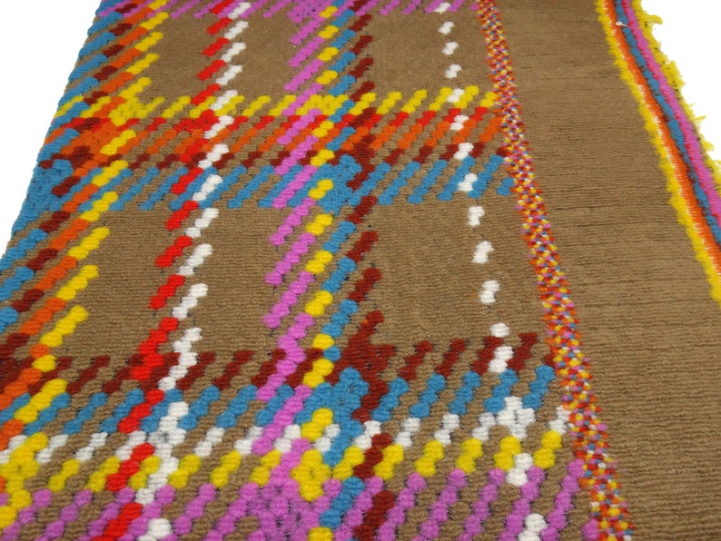 Puede incluir: Primer plano de una alfombra marr&oacute;n con un patr&oacute;n de cuadros de colores. La alfombra est&aacute; hecha de hilo tejido y tiene un borde de flecos.