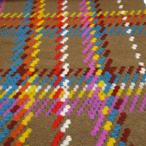 Puede incluir: Una alfombra marr&oacute;n con un patr&oacute;n de cuadros de colores. El patr&oacute;n est&aacute; formado por peque&ntilde;os bucles redondos tejidos en tonos de rojo, naranja, amarillo, azul, rosa y blanco.