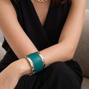 May include: A gold chain necklace and a teal leather cuff bracelet with silver-tone metal accents. The bracelet has a textured surface. The person is wearing a black sleeveless top.