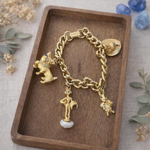 May include: Gold-toned charm bracelet with a chunky chain design. The bracelet features several charms, including a dog, a figure, and a rabbit, all adorned with small blue accents. The bracelet is displayed on a wooden tray.