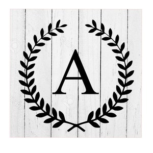 May include: Black and white image of the letter A surrounded by a wreath of leaves on a white wood background.