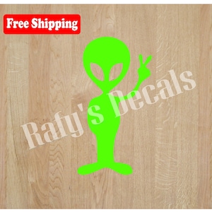 May include: Green alien silhouette with a peace sign, on a wood background. The alien is wearing a peace sign on its hand. The text "Rafi's Decals" is visible in the background.