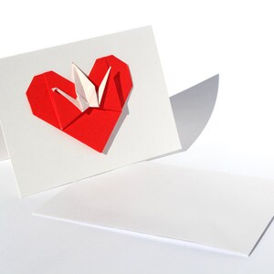 May include: A white greeting card with a red origami heart and a white origami crane. The card is folded, with the heart and crane on the front. A separate, unfolded card lies below.
