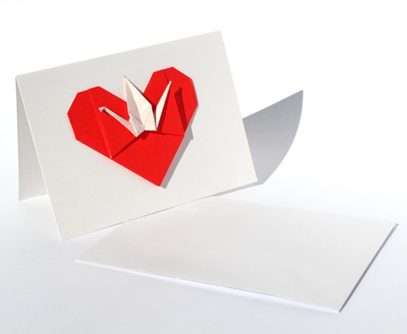 May include: A white greeting card with a red origami heart and a white origami crane. The card is folded, with the heart and crane on the front. A separate, unfolded card lies below.