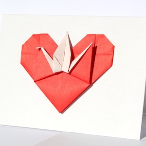 May include: A white greeting card featuring a red origami heart with a white origami crane in the center. The heart is folded from red paper, and the crane is made of white paper. The card is on a white background.