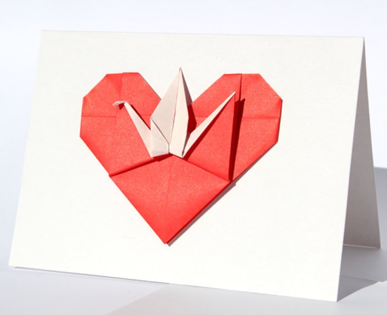May include: A white greeting card featuring a red origami heart with a white origami crane in the center. The heart is folded from red paper, and the crane is made of white paper. The card is on a white background.