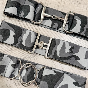 May include: Three equestrian belts with a camouflage pattern in shades of gray and black. Each belt features a different silver-colored buckle design: a snaffle bit, a rectangular buckle, and a round O-ring buckle.