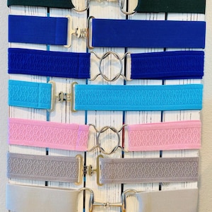 May include: A collection of equestrian belts in various colors and patterns, including navy, green, blue, turquoise, pink, gray, beige, black and white diamond, and red plaid. Each belt features a silver-toned buckle.
