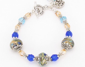Könnte beinhalten: Ein silbernes Armband mit blauen und goldenen Glasperlen, zwei größeren Perlen mit Wirbelmuster und einem silbernen Herzanhänger. Das Armband hat einen Schiebeverschluss.