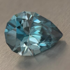Puede incluir: Una gema facetada en forma de pera, con un brillante tono azul claro. Las m&uacute;ltiples facetas de la gema reflejan la luz, creando un efecto brillante. La gema se encuentra sobre un fondo neutro y texturizado.