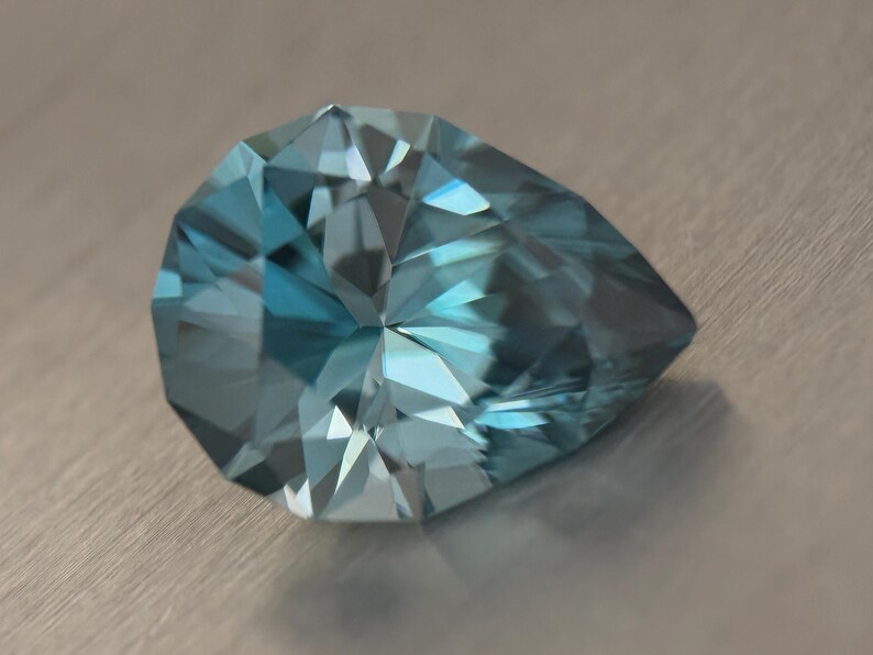 Puede incluir: Una gema facetada en forma de pera, con un brillante tono azul claro. Las m&uacute;ltiples facetas de la gema reflejan la luz, creando un efecto brillante. La gema se encuentra sobre un fondo neutro y texturizado.