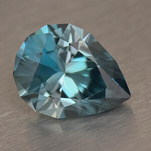 Puede incluir: Una gema facetada en forma de pera con un llamativo tono azul-verdoso. Las m&uacute;ltiples facetas de la gema reflejan la luz, creando un brillo brillante. La gema se encuentra sobre una superficie met&aacute;lica neutra.