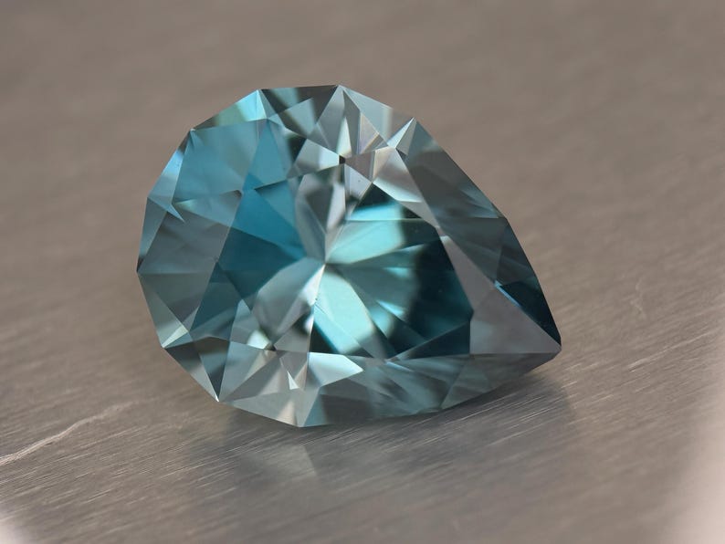 Puede incluir: Una gema facetada en forma de pera con un llamativo tono azul-verdoso. Las m&uacute;ltiples facetas de la gema reflejan la luz, creando un brillo brillante. La gema se encuentra sobre una superficie met&aacute;lica neutra.