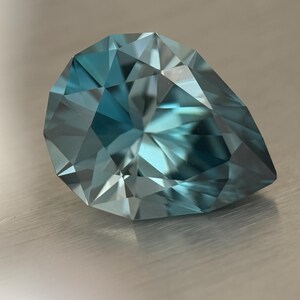 Puede incluir: Una gema facetada en forma de pera, de color azul verdoso. Las m&uacute;ltiples facetas de la gema reflejan la luz, creando un efecto brillante. La piedra se encuentra sobre una superficie met&aacute;lica cepillada.