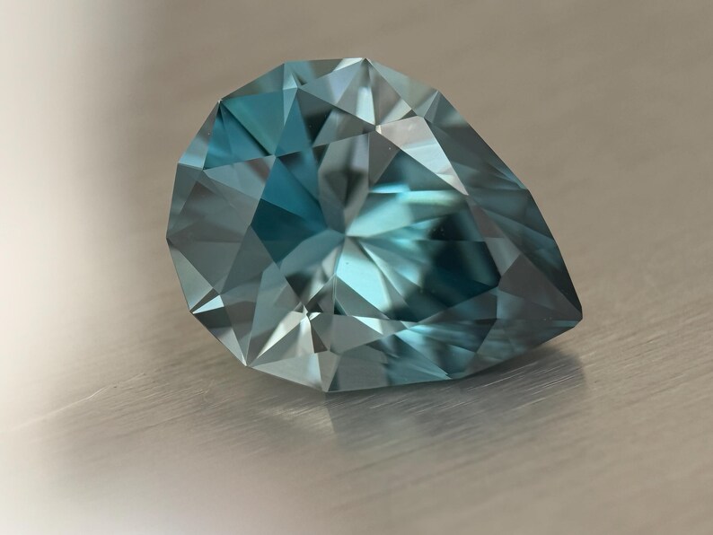 Puede incluir: Una gema facetada en forma de pera, de color azul verdoso. Las m&uacute;ltiples facetas de la gema reflejan la luz, creando un efecto brillante. La piedra se encuentra sobre una superficie met&aacute;lica cepillada.