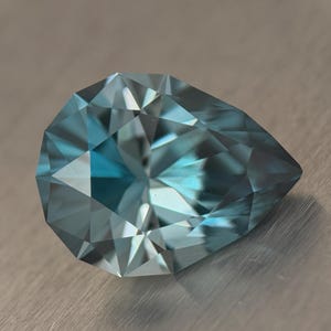 Puede incluir: Una gema facetada en forma de pera, de un tono azul verdoso. La superficie de la gema es un mosaico de facetas triangulares, que capturan y reflejan la luz. La piedra se encuentra sobre un fondo met&aacute;lico neutro, resaltando su brillo.