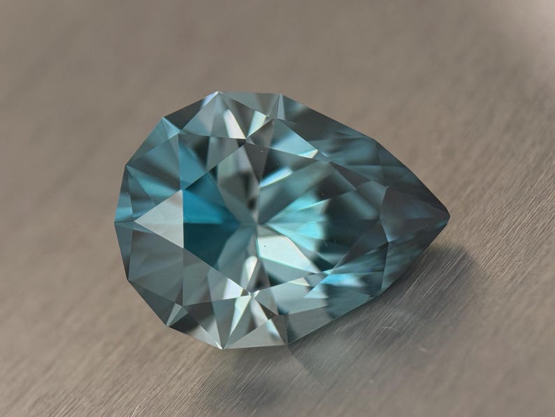 Puede incluir: Una gema facetada en forma de pera, de un tono azul verdoso. La superficie de la gema es un mosaico de facetas triangulares, que capturan y reflejan la luz. La piedra se encuentra sobre un fondo met&aacute;lico neutro, resaltando su brillo.