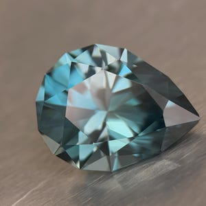 Puede incluir: Una gema facetada en forma de pera con un degradado de color turquesa y azul. La gema tiene muchas facetas que reflejan la luz, creando un efecto brillante. La gema est&aacute; sobre un fondo neutro.
