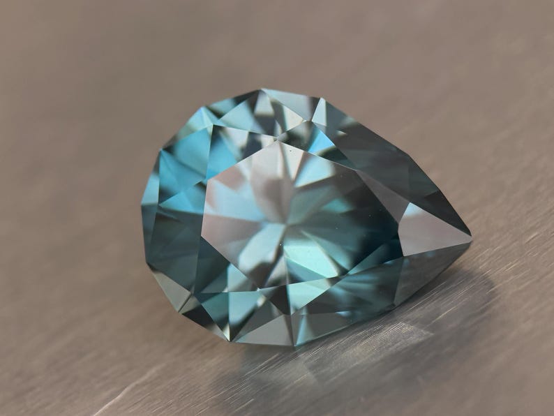 Puede incluir: Una gema facetada en forma de pera con un degradado de color turquesa y azul. La gema tiene muchas facetas que reflejan la luz, creando un efecto brillante. La gema est&aacute; sobre un fondo neutro.